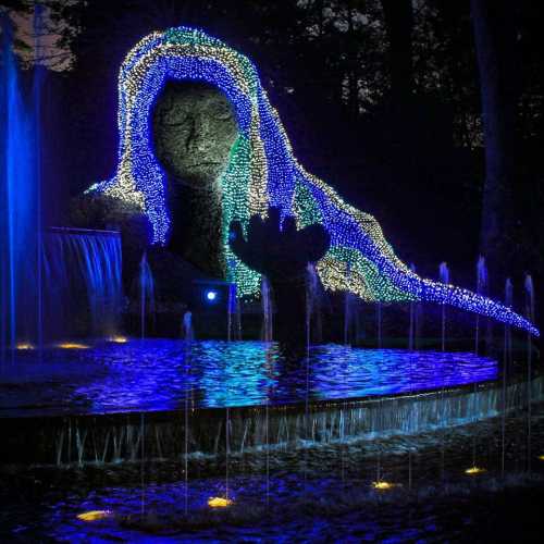 A stone statue illuminated with colorful lights, surrounded by a water feature and trees at night.