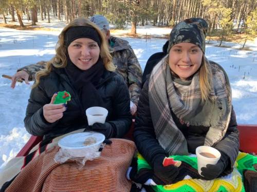 Two women sit on a sled in the snow, holding cups and snacks, wrapped in blankets, with trees in the background.