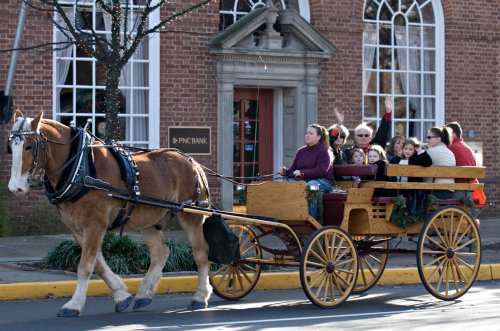A horse-drawn carriage with passengers rides past a brick building, decorated for the holidays.