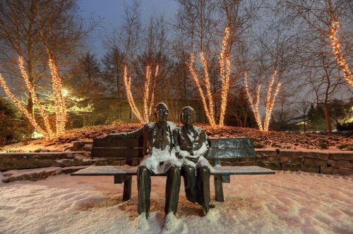 A snow-covered statue of two figures sitting on a bench, surrounded by trees adorned with warm lights at night.