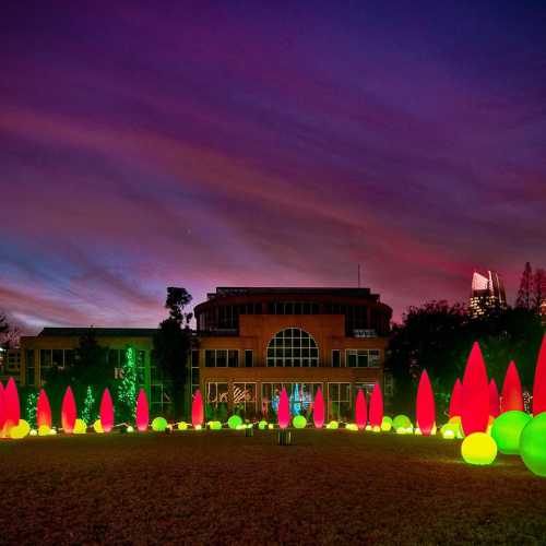 Colorful lights illuminate a park at dusk, with vibrant green and red decorations against a dramatic sky.