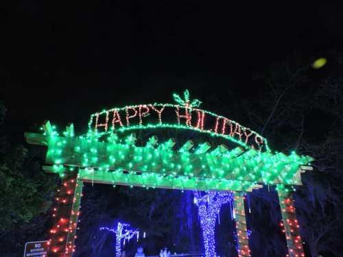 A festive display of colorful holiday lights spelling "Happy Holidays" on a wooden arch at night.