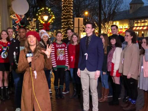 A group of people in festive attire stands outdoors, while a woman interviews someone with a microphone. Holiday decorations are visible.