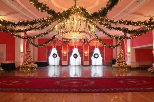 Elegant banquet hall decorated for Christmas with garlands, wreaths, and trees, featuring a grand chandelier and polished floor.