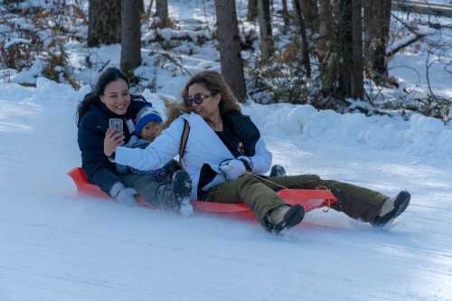 Three people sledding down a snowy hill, smiling and enjoying the winter day. Trees are visible in the background.