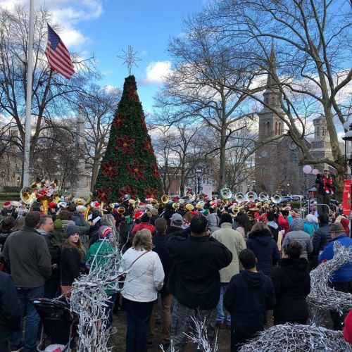 A crowd gathers around a decorated Christmas tree, with musicians performing in a festive outdoor setting.