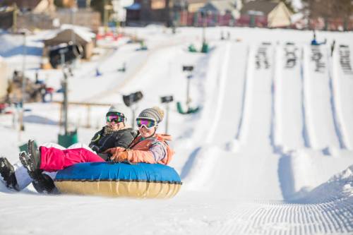 Two people sit on a snow tube, smiling as they slide down a snowy hill with other tubing lanes in the background.
