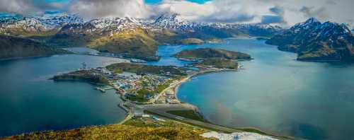 Aerial view of a coastal town surrounded by mountains and water, with snow-capped peaks in the background.