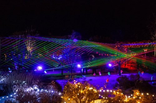 Colorful laser lights create vibrant patterns in a dark outdoor setting, surrounded by festive decorations.