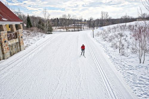 A lone skier glides on a snowy trail beside a rustic building and trees under a cloudy sky.