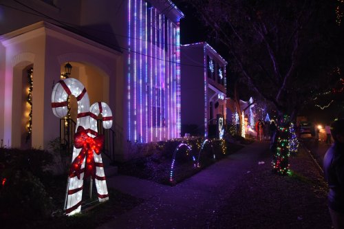 Colorful holiday lights decorate houses along a sidewalk, featuring candy cane displays and festive decorations.