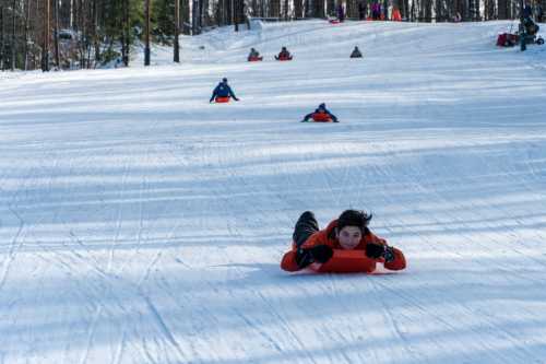 A person in an orange jacket sleds down a snowy hill, with others in the background enjoying the winter activity.