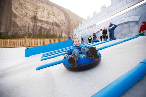 A child joyfully sleds down a snowy slope on a blue tube, surrounded by a winter-themed park and other visitors.