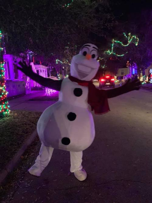 A person in a snowman costume with a scarf poses cheerfully on a decorated street at night.