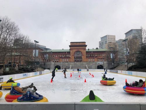 People ice skating on a rink with colorful inflatable tubes, surrounded by buildings and trees on a cloudy day.