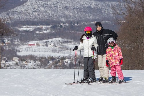 A family of three stands on a snowy slope, ready to ski, with a scenic winter landscape in the background.