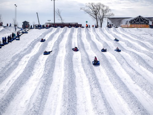 A snowy hill with people sledding down on tubes, surrounded by trees and a cloudy sky.