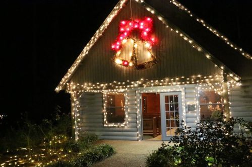 A cozy cabin decorated with festive lights and a large red bow, creating a warm holiday atmosphere at night.