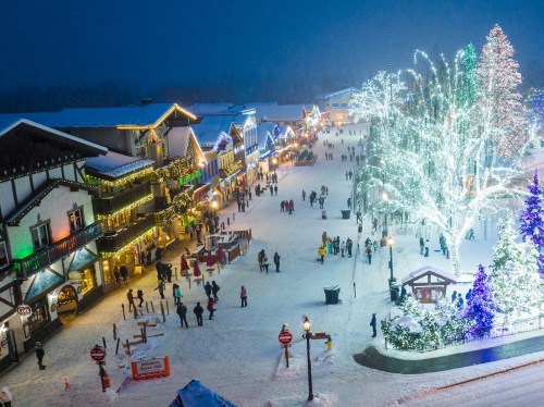 A snowy street scene with festive lights, decorated trees, and people enjoying a winter celebration.