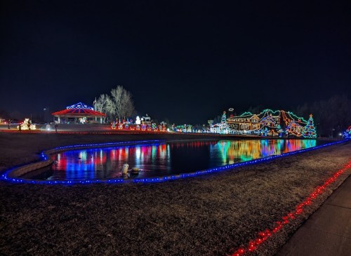 Colorful holiday lights reflect on a pond at night, with decorated trees and festive displays in the background.