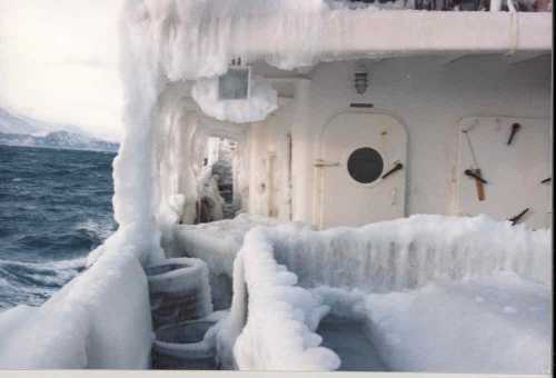 A ship's deck covered in ice, with frozen railings and a view of turbulent sea in the background.