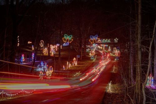 A festive display of colorful holiday lights along a dark road, with trees and decorations visible in the background.