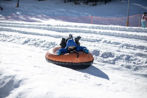 A child in a blue jacket sleds down a snowy hill on an orange inflatable tube. Snowy landscape in the background.