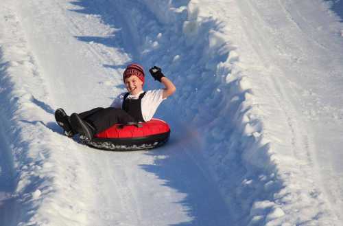 A child in a red snow tube joyfully slides down a snowy hill, raising one hand in excitement.