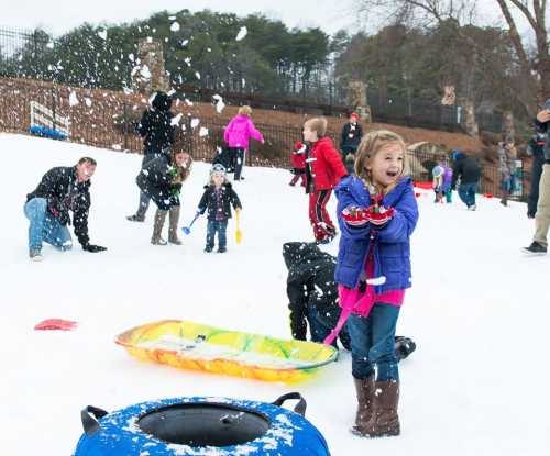 A joyful girl in winter clothes plays in the snow, surrounded by families tubing and enjoying a snowy day.