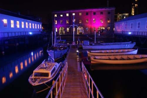 A nighttime view of a dock with boats and a brightly lit building reflecting in the water.