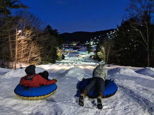 Two people sit on snow tubes at night, overlooking a snowy hill with lights and a tubing course in the distance.