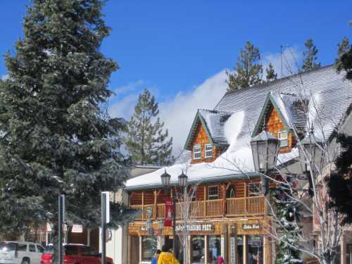 A snowy scene featuring a charming wooden building with a steep roof, surrounded by tall evergreen trees and blue skies.