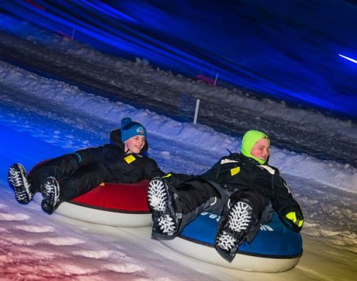 Two children in winter gear sliding down a snowy hill on colorful snow tubes, with blue and purple lights in the background.