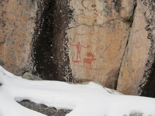 Ancient rock art depicting figures and symbols on a stone surface, partially covered by snow.