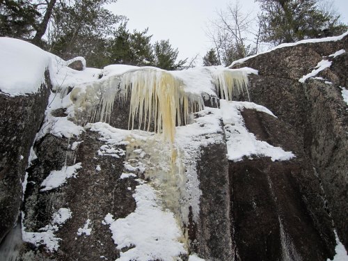 Icicles hang from a rocky cliff, surrounded by snow and trees, creating a winter landscape.