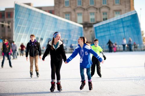 Two girls skate hand-in-hand on an ice rink, smiling, with others skating in the background and a modern building behind them.