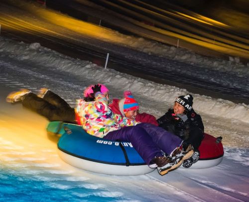 A group of four people in colorful winter clothing enjoying snow tubing at night on a snowy slope.