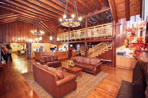 Cozy lodge interior with wooden beams, leather sofas, a coffee table, and a staircase leading to a second level.