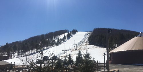 A snowy ski slope under a clear blue sky, with trees and a building in the foreground.