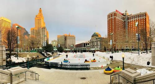 A winter scene featuring an ice rink surrounded by buildings, with people skating and colorful bumper cars on the ice.
