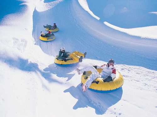 Four people in yellow snow tubes sliding down a snowy hill on a sunny day.