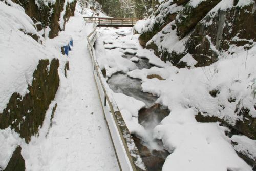 A snowy landscape with a flowing stream and a wooden bridge crossing over it, surrounded by rocky cliffs and trees.