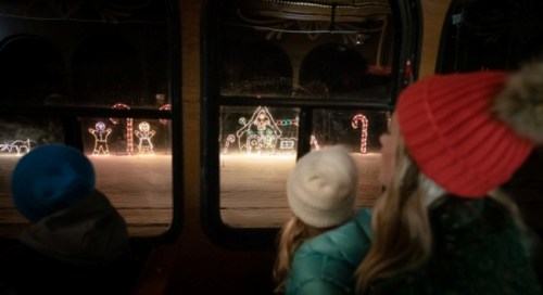 A woman and two children watch colorful holiday lights from a train window, with festive decorations in view.