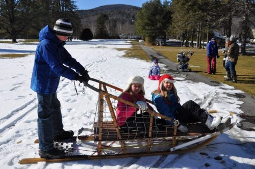 A boy pulls a wooden sled with two girls sitting on it, surrounded by snow and trees on a sunny winter day.