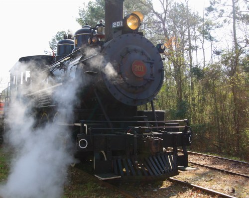 A vintage steam locomotive with smoke billowing, set against a backdrop of trees and sunlight.