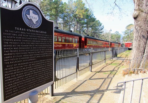 Historic Texas State Railroad with red train cars alongside a black historical marker and trees in the background.