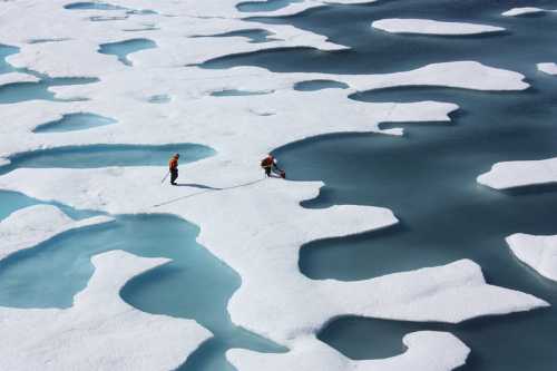 Two researchers walk on melting ice with water pools, highlighting climate change effects in a polar region.