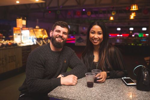 A smiling man and woman sit at a table with drinks in a lively indoor setting.