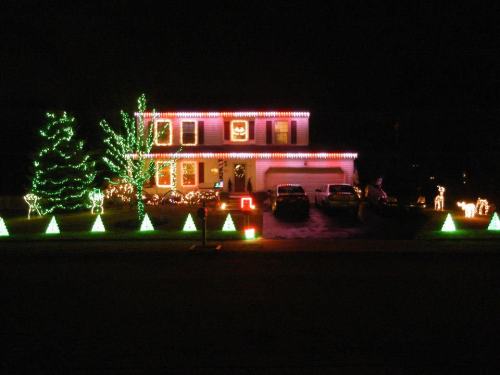A house decorated with colorful Christmas lights, featuring trees and festive displays in the yard at night.