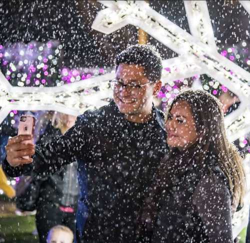 A couple takes a selfie in a snowy setting, surrounded by colorful lights and festive decorations.